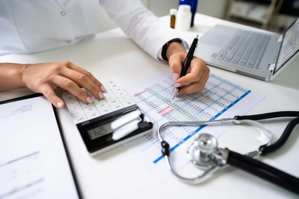 Person using a calculator and writing on a medical spreadsheet with a stethoscope and laptop nearby.