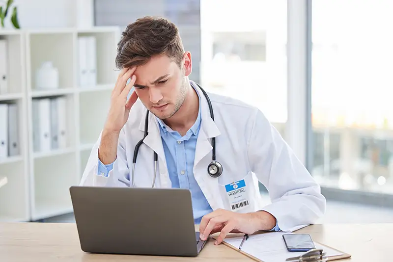 A doctor stressed while working on a laptop, likely dealing with billing issues.
