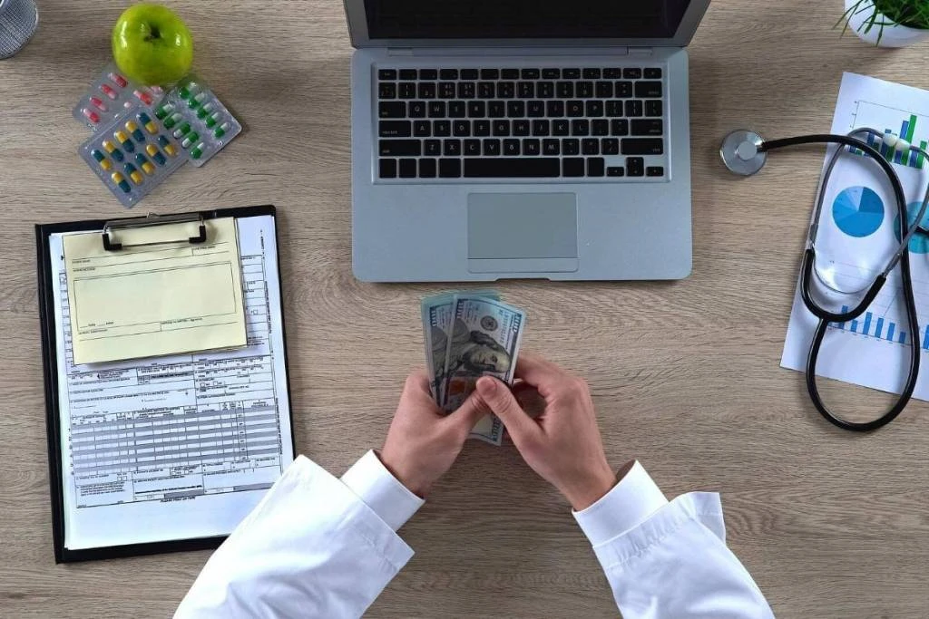 Doctor reviewing medical charts and calculating billing costs with a laptop and stethoscope nearby