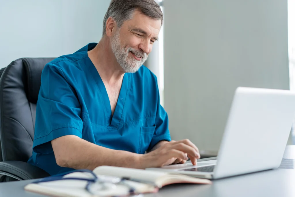 A doctor in blue scrubs smiling while typing on a laptop, with a stethoscope and notebook on the desk.