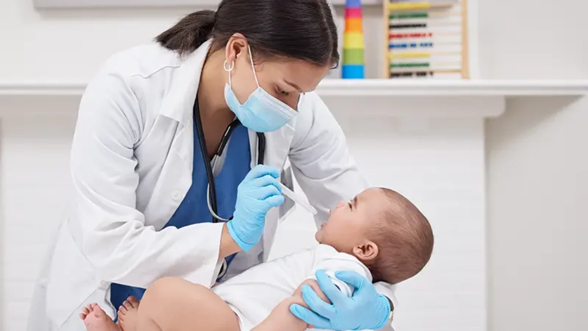 A doctor wearing a mask and gloves taking a temperature of a baby during a check-up.