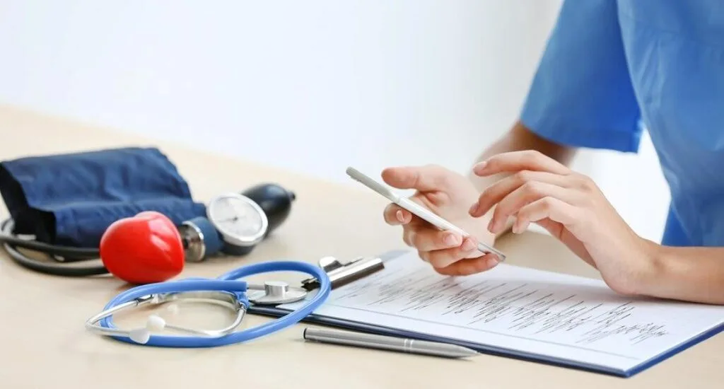 A healthcare professional checking their phone while sitting at a desk with a stethoscope and blood pressure cuff.