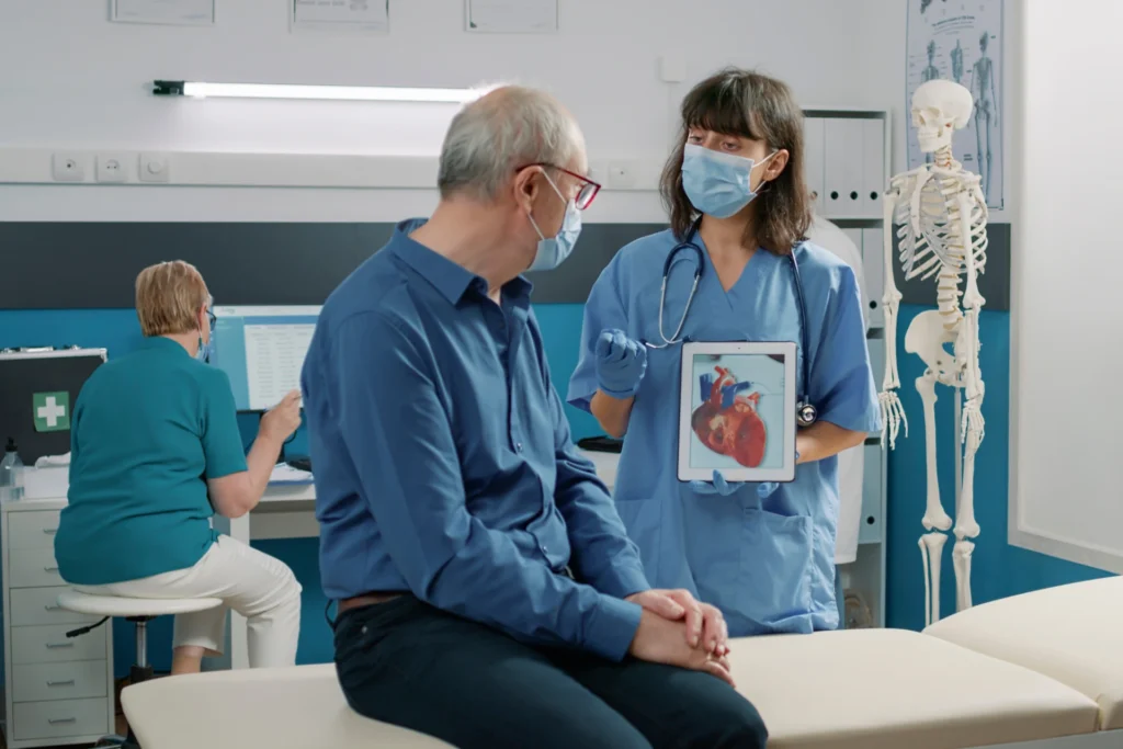 A healthcare professional showing a tablet with a heart model to a patient while another staff member works at a computer