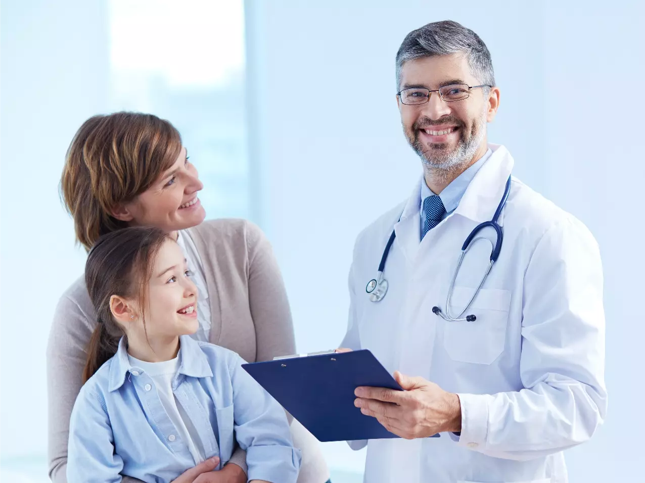 A doctor smiling with a child and parent while holding a clipboard.