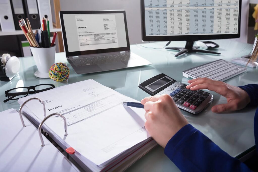 The image shows a person reviewing invoices and using a calculator, with a laptop displaying a spreadsheet in the background.