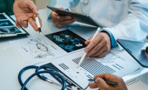 The image shows medical professionals collaborating over documents with digital devices, discussing medical data and illustrations.