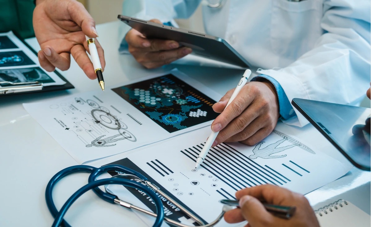 The image shows medical professionals collaborating over documents with digital devices, discussing medical data and illustrations.