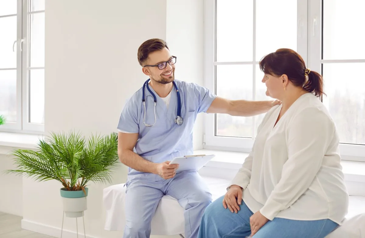 A doctor in scrubs is offering reassurance to a patient sitting on an exam table in a bright medical office, with a friendly and supportive gesture.