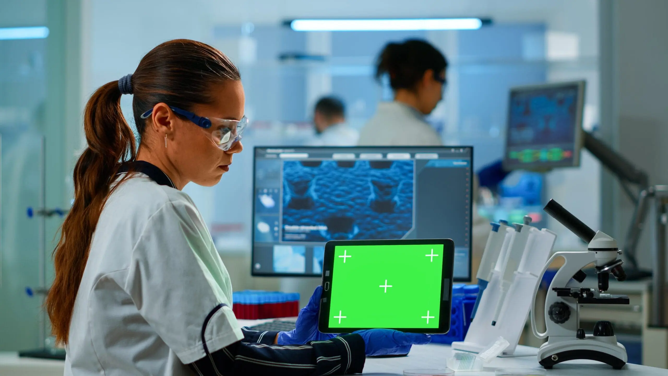 A lab technician holding a tablet with a green screen in a laboratory setting.