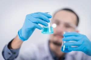 A laboratory technician wearing blue gloves holding two glass beakers with blue liquid.