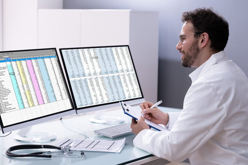A medical professional reviewing spreadsheets on dual monitors while taking notes.