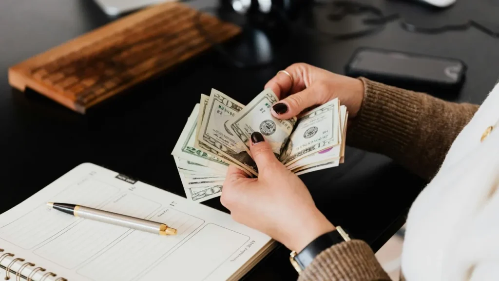 A person counting stacks of cash with a pen and notebook on the desk.