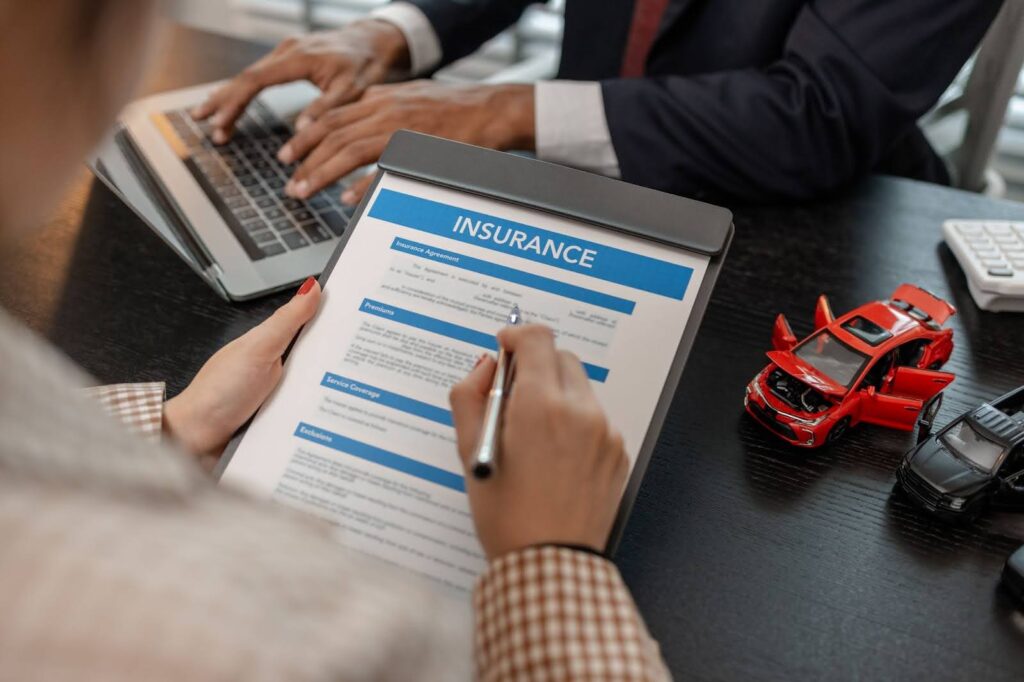 A person is reviewing a car insurance document while a laptop and toy cars are visible on the table.