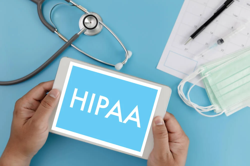 Hands holding a tablet displaying the word 'HIPAA' with medical items in the background.