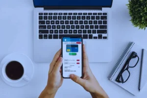 The image shows a person using a smartphone with a digital payment or banking app open, alongside a laptop, coffee cup, and other items on a desk.