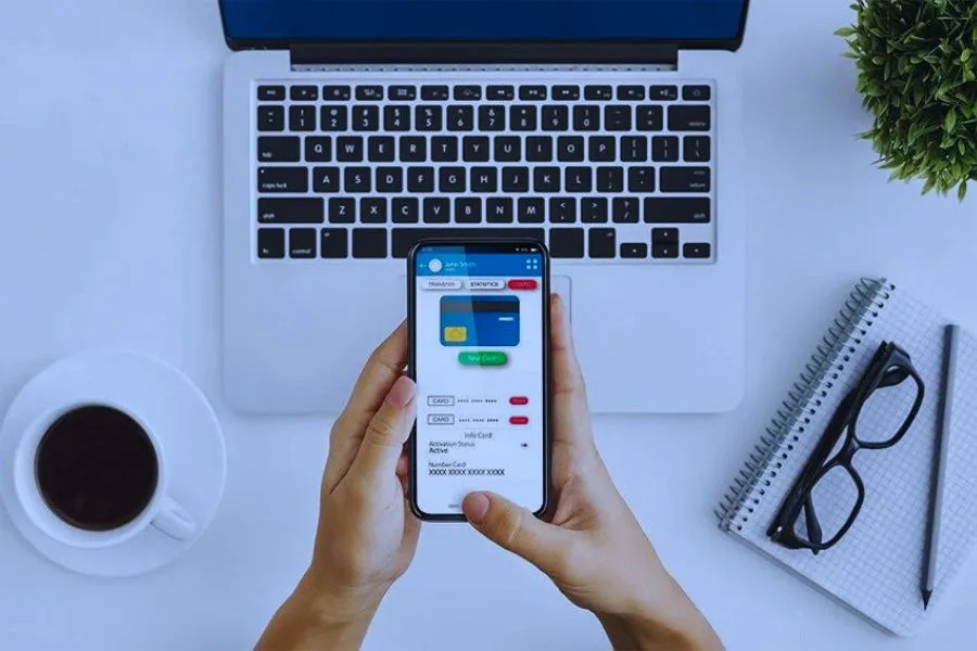 The image shows a person using a smartphone with a digital payment or banking app open, alongside a laptop, coffee cup, and other items on a desk.