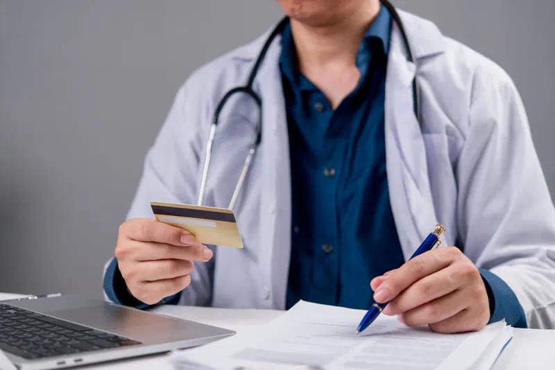 a healthcare professional holding a credit card in one hand and a pen in the other, while reviewing documents at a desk with a laptop for eft in medical billing