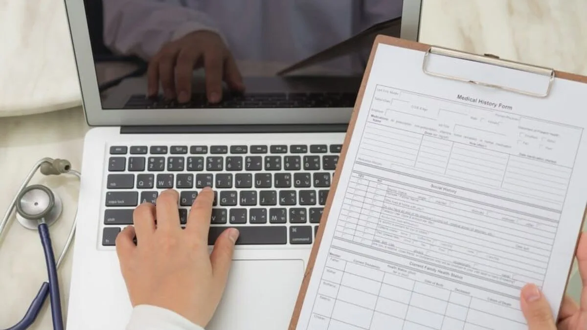 a person typing on a laptop while holding a medical history form on a clipboard.